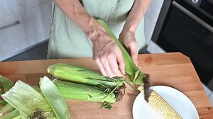 Download woman cleans fresh corn from green leaves in the kitchen at home. Home cooking, organic sweet yellow corn, healthy vegetable season. Process of cleaning corn cobs for cooking for free
