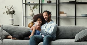 Smiling Black father adult older brother relax on couch cuddle tween girl daughter younger sister hold phone watch video laugh with kid. Dad schoolgirl friends enjoy funny online activity using cell