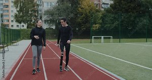 Joyful man with prosthetic running blade chatting with positive woman. Male athlete with amputed leg and happy female person communicating and smiling while walking at sports field.