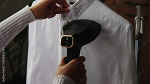 woman's hand uses a garment steamer, smoothing out wrinkles on a shirt with the steamer, releasing a stream of hot steam. Steam iron in the home