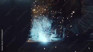 Skilled professional worker in a protective mask performing metal inert gas welding, with bright sparks and smoke flying from the hot metal arc in a dark industrial workshop