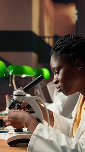 Vertical Dedicated female student working with a microscope to prepare for microbiology lessons in a library, focusing on microorganisms and chemical processes. Notes for her academic project. Camera A.