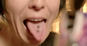 Body modification tongue piercing. Woman face shows into camera phone, smartphone bokeh. Two earrings, she moves, covers her mouth, smiles. Metal earrings straight barbells with metal beads.