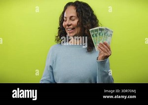 Middle-aged woman holding israeli shekel banknotes against a vibrant yellow background, smiling while extending her hand, conveying a sense of wealth and financial happiness.