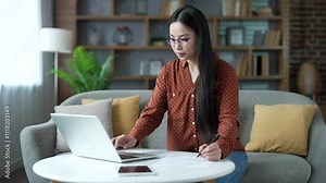 Confident young Asian woman dealing with household finances using laptop computer sitting on sofa in home office. Busy female calculates household budget, takes notes, fills out the form of documents