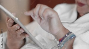 Senior woman lying on a bed in a white terry robe uses a tablet, surfs Internet sites. Slow Motion, close-up.