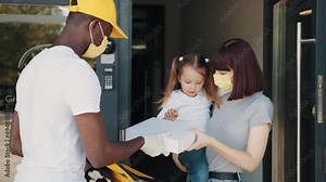 Home delivery. An African American courier hands a box to a girl with a child in a mask. Young courier with mailbox with package, customer receives parcel during curfew, coronavirus pandemic.