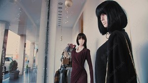 The mannequins are fair-skinned women with black hair. Storefront display. Two mannequins behind glass in the store wearing beautiful dresses and carrying shoulder bags.