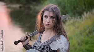 Medieval warrior woman with war paint on her face, dressed in chain mail armor with a sword and a shield in her hands, posing standing against the backdrop of bokeh sunset, river and forest.