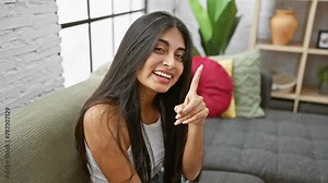 Excited young indian woman sitting on the sofa, pointing a finger up with a happy face, having a sudden smart idea at home. relax and think one question in mind, showing number one with joy.