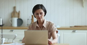 Attractive serious middle-aged 55s woman working at home using laptop, prepare document, makes on-line payment through e-bank application looks focused sit at table in domestic kitchen. Telework, tech
