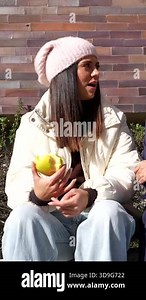 Two stylish young women in winter clothes chatting together on a sunny day while sitting on a bench against a brick wall, each enjoying a healthy apple snack during their conversation Stock Video Footage - Alamy