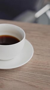 White cup of black coffee on the wooden table. Female hands pull the cup closer. Laptop and phone are on the desk beside. Close up. Vertical