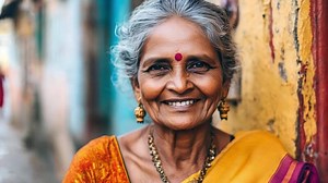 Download An elderly indian woman with a broad smile wears a beautifully colored sari and traditional jewelry, standing against a backdrop of brightly painted walls in an urban area. for free