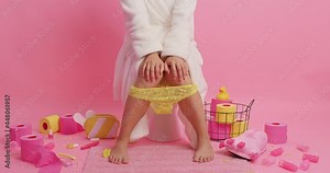Unrecognizable woman wears white soft bathrobe lacy yellow pants pulled down on legs keeps hands on knees reveals herself sits on toilet bowl isolated over pink background. Domestic routine.