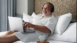 A man on vacation attends a video conference in a bathrobe, sitting on the bed with headphones and a laptop. A casual yet professional scene, blending relaxation and remote work.