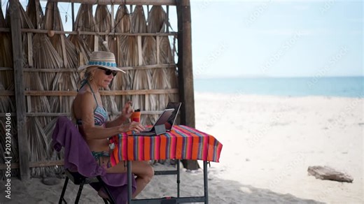 Mature woman has happy video chat on computer tablet, while on a beach during vacation time in a tropical setting.