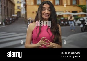 Woman in pink tank top clenches fists under tight grin on urban street lined with scooters and amber buildings; nervousness.