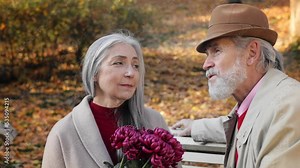 Happy old mature retired couple sitting on park bench, talking and smiling to each other. Middle aged spouses having a date, enjoying time together in beautiful autumn park.