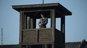 Barb wire fence. A young German soldier with a rifle standing in a watchtower and looking around. Bottom view. World War 2 reenactment