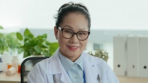 Tilt up of successful mature doctor typing on laptop keypad while consulting online patient and then looking at camera with smile while sitting by workplace in medical office