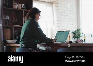 Adult woman wearing glasses working on a laptop while sitting in a bright kitchen with panoramic windows in a country house. Remote work, home office and modern lifestyle concept Stock Video Footage - Alamy