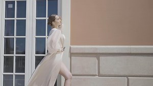 Side view of a gorgeous female model in a silk dress swaying in the wind. Action. Woman standing and posing in front of a building with big window and brown facade.