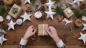 Aerial lockdown footage of woman tying ribbon on wrapped gift box at wooden table. Overhead flat lay of Christmas gifts and ornaments surrounding female. Lady is preparing for festive season.