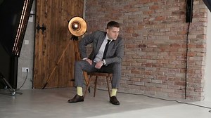 A fashionable brutal guy in a gray suit sits on a chair in the Studio and poses for a photographer against a brick wall and an old lamp.