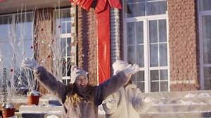Playful girlfriends throwing up snow on decorated countryside cottage background. Happy teenager girls throwing up fluffy snow on Christmas walk. Positive people emotions on New Year eve