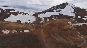 Aerial view of Icelandic mountains with snow, vibrant orange and brown hues, winding paths, and eroded geothermal formations in motion.