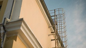 House with iron safety ladder on bright yellow facade under clear sky, featuring rooftop access structure, modern exterior design, and metal framework for safety, convenience, and architectural