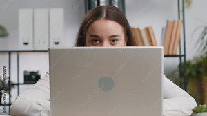 Confident business woman hiding behind laptop computer, looking at camera, spying his colleagues working, peeping. Professional freelancer girl looking from behind computer with cunning eyes glance