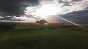 Aerial view of a solitary mountain in Iceland, surrounded by flatlands and winding rivers. The camera sweeps slowly under sunlight breaking through clouds.