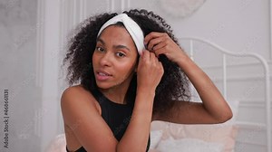 Dark skin woman putting on headband and smiling at camera, in bright room. Young black girl doing her morning routine. Morning beauty routine of black woman. High quality 4k footage