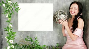 beautiful smiling woman posing in spring photo session holding cute white rabbit in wicker basket, green plant and flower decoration of photo studio, free space for text, white background for letters