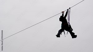 Instructor and schoolchild ride zip-line in winter cable park against gray cloudy sky. Worker coming to help frightened child on ropes