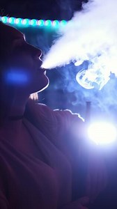 Close up side view of a girl smoking hookah on blue neon background. Media. Female while smoking a hookah in a club, exhaling a fog cloud and chilling.