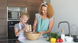 Pretty female housewife cooker wears apron and headband on head, has glad cheerful expression while demonstrating little blond girl how to make cake, Home Cooking, Homemade Food