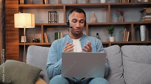 Friendly teacher wearing wireless headset sitting on sofa doing distance learning online chat via video conference call, communication laptop webcam. Home remote education, english lessons at home.