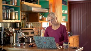 Mature woman working from home on laptop computer in home kitchen as camera slides to the left.