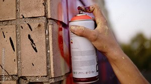 Close-up of artists hand dirty in paint applying spray drawing a red coloured line on a street building bricked wall. Action. Stained fingers of artist hold spray can with colored paint on concrete