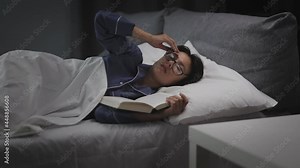 Tired middle aged woman falling asleep while reading book in bed. Caucasiana brunette in eyeglasses ad pajamas lying in bed with soft pillows and blanket. Stock Video