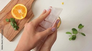 Woman taking out freezed ice cubes and putting them into a glass with orange pulp and mint inside, dropping a few cubicles on the white table. Cooking a simple recipe lemonade on a hot summer day.