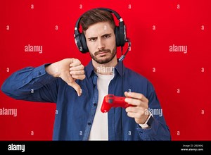 Hispanic man with beard playing video game holding controller with angry face, negative sign showing dislike with thumbs down, rejection concept Stock Photo - Alamy