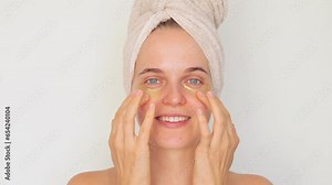 Beauty and skincare regimen. Cosmetic facial treatment. Smiling delighted woman wearing bath towel carefully places under-eye patches on her face skincare procedure isolated over white background.