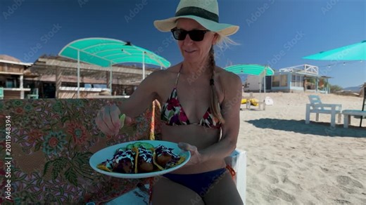 Mature woman sits at beach taco stand and eats delicious shrimp tacos while enjoying the sun and view at Playa Blanca in Baja California