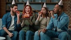 Friends sing happy birthday song to woman congratulate girl at home. Diverse ethnic teammates women and men in party caps presenting festive cake with candles celebrating birth event blowing whistles