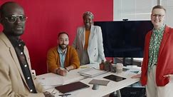 Full zoom-out shot of team of four creative multiracial male and female team members posing together in bright, stylish office against red wall, smiling confidently, while having project meeting