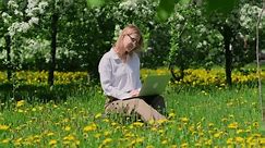 Attractive adult woman is sitting on a green lawn in a park with a laptop on a sunny summer day. Happy female freelancer works among flowering trees illuminated by sun.